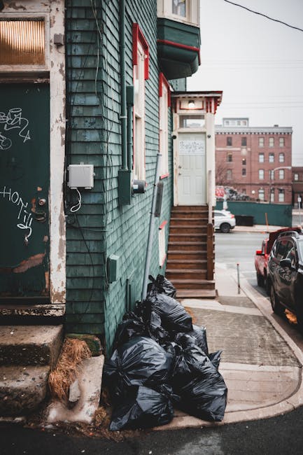 The image depicts the exterior corner of a building with green wooden cladding and red-bordered white window frames, situated on an urban street. A staircase with wooden steps and a white door at the top leads to a raised entrance. Several black rubbish bags are piled against the building's side near the steps, indicating waste collection for private disposal or alternative waste handling, as often managed by private rubbish removal services like Rubbish Removal Kennington. Adjacent to the bags, there are concrete steps leading down to a small sidewalk area, with some dirt or debris accumulated at the base. To the left, part of a garage door or shutter with graffiti tags is partially visible, showing a rusted or weathered metal surface. In the background, parked cars and a brick building with multiple windows are visible, under an overcast sky that softens the overall lighting, emphasizing the everyday urban environment and the need for effective rubbish removal solutions in the area.