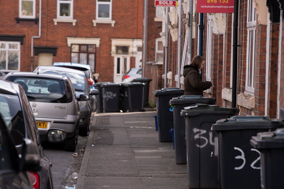 A residential street scene showing a row of dark grey wheelie bins placed along a sidewalk in front of red brick houses with white window frames. The bins are lined up closely, some with open lids, and are marked with white spray-painted numbers. A woman wearing a brown jacket and facing away from the camera is seen near the bins, appearing to be either opening or closing one of them. Parked cars, including a silver hatchback and other vehicles, are situated along the road, which has a grey concrete pavement and a street surface showing patches of wear. The background reveals additional residential buildings, some with small front gardens or driveways, and a partially visible red sign with white text hanging from one of the houses. The overall environment depicts typical urban residential waste management, with spaces allocated for rubbish collection and private waste handling, aligning with independent rubbish removal services in the area.