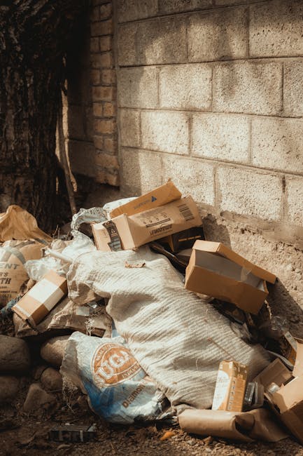 A pile of discarded cardboard boxes, some open and flattened, and paper packaging materials are leant against a rough brick wall outdoors, next to a tree trunk. The boxes vary in size and have printed labels and barcode markings visible on their surfaces. Some of the boxes are partially torn or crumpled, lying on a ground surface composed of dirt and scattered small rocks. A large beige sack made of woven material is positioned among the waste, containing additional debris. The scene appears to depict the accumulation of bulk rubbish intended for collection or clearance, characteristic of private disposal or on-site rubbish removal. The brick wall serves as a backdrop, with natural light illuminating the scene, highlighting the textures of the cardboard, paper, and ground. This outdoor setting supports the concept of independent rubbish handling, aligning with services offered by companies such as Rubbish Removal Kennington, as they manage waste at such locations.