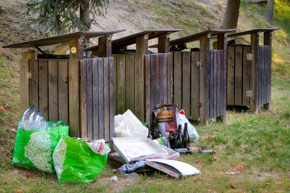 A series of four wooden outdoor rubbish bins with small slanted roofs, positioned on a grassy patch beside a sloped, wooded area with trees and foliage in the background. The bins are made of vertical wooden planks, stained dark brown, with metal hinges and locks on the front. Some of the bins have visible weathering and slight warping. In front of the bins, there are various waste items including a large green reusable shopping bag filled with empty plastic bottles, a white pizza box, a black plastic bag, and several glass bottles and cans, some of which are partially collapsed or tipped over. The ground is a combination of grass and dirt with scattered fallen leaves. The scene appears to be an informal waste disposal area, illustrating a potential issue for private rubbish collection services like those offered by Rubbish Removal Kennington, especially in the context of inaccessible or unmanaged communal waste points in residential estates or outdoor communal spaces.