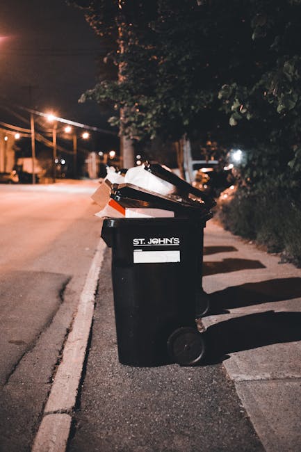 A black wheeled trash bin labeled 'St. John's' positioned on a residential pavement at night, with its lid open revealing discarded waste. The surrounding environment includes a quiet street illuminated by streetlights, with a tree partially visible overhanging the sidewalk and neighboring houses or buildings faintly lit in the background. The bin appears to contain various rubbish, including paper and plastic items, which are somewhat chaotically stacked, emphasizing the need for proper waste management or alternative rubbish removal services. Shadows cast by streetlights stretch across the pavement, creating a calm, dark atmosphere reflective of urban residential areas. The scene is relevant to discussions about waste disposal and rubbish collection, illustrating typical household waste awaiting collection by private waste removal companies, such as Rubbish Removal Kennington, supporting environmentally responsible rubbish disposal practices.