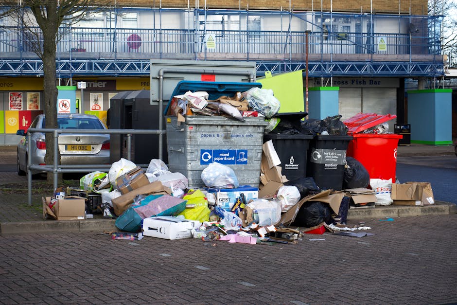 A large collection of overflowing waste bins and scattered rubbish bags, cartons, cardboard boxes, paper, and plastic packaging piled on a paved area adjacent to a parking space. The waste includes black, blue, and red refuse containers, some of which have open lids revealing mixed garbage contents. The black and blue bins have labels indicating they are for mixed paper and cardboard disposal, while the red bin appears to be for general waste. Surrounding the bins are loose piles of discarded materials, some spilling onto the ground, including flattened cardboard boxes, plastic bags, and assorted packaging debris. In the background, a building with a blue construction scaffold and façade, along with a row of shopfront windows and signage, can be seen, indicating a commercial area. A parked car is situated behind a metal railing, and a tree is partially visible on the left side. The scene suggests an instance of improperly managed waste collection, possibly related to independent rubbish disposal services, aligning with private waste removal themes within a town or urban environment.
