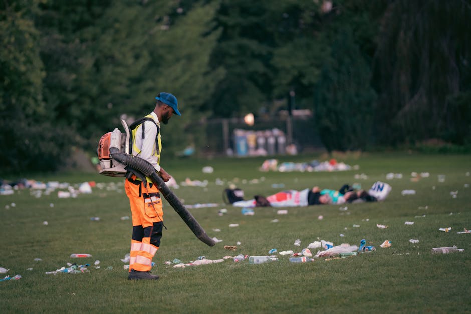 A street cleaner wearing a blue cap, high-visibility orange trousers with reflective strips, and a white jacket is using a leaf blower or vacuum to remove litter from a grassy park area. The worker carries a backpack-style collection device and is focused on clearing the scattered debris, which includes plastic bottles, cups, and paper, spread across the green lawn. In the background, two individuals are lying on the grass amidst more rubbish, with trees and a small waste disposal unit visible beyond them. The scene appears to be either early morning or late evening, with soft natural lighting emphasizing the contrast between the clean-up activity and the scattered mess. This image illustrates the process of private waste handling in publicly accessible green spaces, highlighting the importance of rubbish removal services like those offered by Rubbish Removal Kennington for maintaining cleanliness in community areas used for recreation and leisure activities.