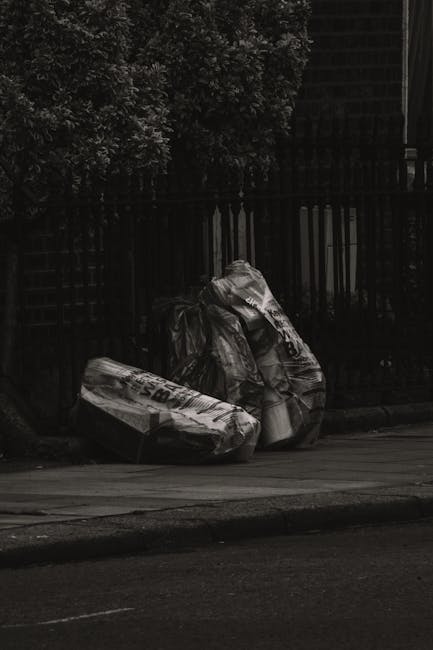 Three large black plastic trash bags filled with bulky waste are positioned on the edge of a paved sidewalk in front of a black metal fence with vertical bars. The bags appear to contain miscellaneous refuse, possibly packaging or household items, and are slightly crumpled and tied at the top. Behind the fence, dense foliage of a leafy shrub or small tree provides some greenery contrast. To the right, a section of building is visible, with what appears to be a dark, possibly brick exterior. The scene is outdoors, illuminated by natural daylight, with shadows cast on the ground. This image reflects an instance of private waste management or rubbish removal, typical of discreet, on-site clearance services provided by providers like Rubbish Removal Kennington, catering to the removal of bulky waste from residential or commercial properties in the area.