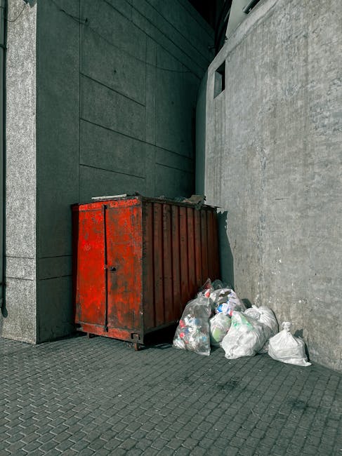 A large collection of mixed household waste is piled on a paved area in front of a commercial building, with multiple black, green, and red wheelie bins, some overflowing with rubbish. The waste includes flattened cardboard boxes, crumpled paper, plastic packaging, and discarded packaging materials. Several paper and cardboard boxes are scattered among the pile, with some partially collapsed or torn. The containers are positioned close together, with the overflowing bin in the center, its lid open to reveal loose rubbish spilling over. Behind the bins, the building façade features a row of shopfronts with closed shutters, and above, there is scaffolding indicating construction work or maintenance, with protective netting visible. A tree with bare branches stands on the left side, and a parked silver car is partially visible behind a metal railing on the left. The scene appears to be set during daylight hours under overcast conditions, highlighting the cluttered nature of the waste collection area, typical of private waste collection or alternative rubbish removal services in an urban environment associated with rubbish removal Kennington.