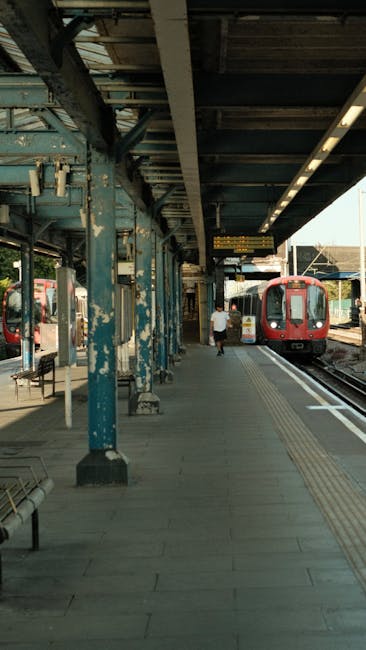 The image shows a train station platform with a red passenger train approaching on the right side. The platform is covered by a weathered metal canopy supported by blue painted steel columns, some of which have peeling paint revealing rust underneath. The surface of the platform is made of grey concrete tiles, with tactile paving running parallel to the train tracks for accessibility. A few benches are visible, including one toward the left side of the image, possibly made of metal with wooden slats. In the background, a single passenger wearing casual clothing, such as a white shirt and dark trousers, is walking away from the camera toward the train. The environment appears clean and well-maintained, with lighting mostly natural, enhanced by daylight. The scene captures a typical station platform that could be part of an independent or private railway service, fitting the context of alternative or scheduled public transport. Rubbish Removal Kennington might handle waste from station maintenance or related services, subtly linked to this setting, with no visible waste or rubbish, but the scene emphasizes structured, accessible urban transit infrastructure.