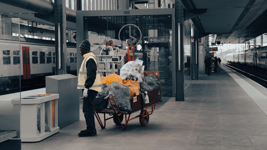 At a modern railway station platform, a worker dressed in dark clothing, a beanie, and high-visibility vest stands beside a hand truck loaded with various bags and loose garbage, including black, grey, and orange plastic trash bags as well as discarded cardboard and paper waste. The bags appear full and are piled on the cart, indicating a collection of waste for removal. Behind the worker, there is a glass-enclosed shelter with metal framework, equipped with cleaning machinery or equipment, and a large circular logo or symbol displayed on the glass. To the left, a white waste bin with a lid is positioned on the platform. In the background, train carriages with closed doors sit on adjacent tracks, and a few other individuals are visible walking or waiting further down the platform. The environment is characterized by sleek, modern architecture with smooth tiled flooring and metallic structural elements, reflecting an organized space where external waste collections, such as those offered by Rubbish Removal Kennington, could be part of alternative waste handling arrangements at busy public transport hubs.
