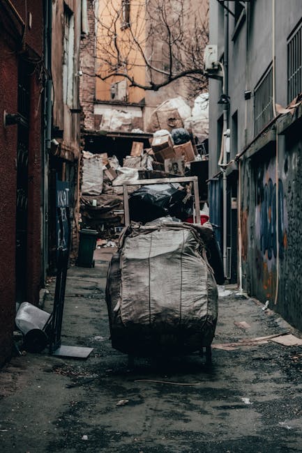A cluttered alleyway in an urban setting, filled with accumulated rubbish and discarded waste materials. In the foreground, a large, grey, fabric refuse bag is positioned on the uneven pavement, which appears stained and weathered. Behind it, a black plastic bin and a small white object are visible on the ground. The alley is flanked by tall, narrow buildings with exterior walls featuring graffiti and graffiti tags, some with metal window grilles and electrical fixtures. The background shows a heap of cardboard boxes and white sacks piled against a wooden barrier, partially covered by a transparent plastic sheet, indicating ongoing rubbish accumulation. Bare tree branches are seen overhead, suggesting the scene occurs during winter or early spring. Diffused natural lighting illuminates the scene, highlighting the textures of the waste and urban environment. This image reflects a typical private waste handling scenario in an alley, emphasizing the importance of regular rubbish removal services, such as those offered by Rubbish Removal Kennington, for maintaining cleanliness and compliance with local disposal regulations.