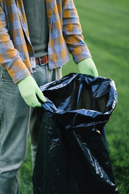 A person wearing a plaid shirt with shades of yellow, blue, and gray, along with green gloves, is holding an open black rubbish sack, which appears to be made of plastic and is partially lined with a silver inner layer. The individual is standing outdoors on a well-maintained grassy area with the background showing a blurred, lush green field. The person's lower body is visible, dressed in gray trousers and a brown belt. The scene suggests an activity related to waste collection or rubbish removal, possibly in a private or community setting, aligning with alternative waste handling practices that may involve personal disposal or onsite clearance. Rubbish Removal Kennington often supports such activities by providing professional waste management services that facilitate responsible rubbish disposal, whether through collection or on-site clearance, in accordance with local regulations or alternative disposal options. The image emphasizes the importance of proper rubbish handling, highlighting the use of appropriate gloves and secure bags to maintain hygiene during waste disposal tasks.