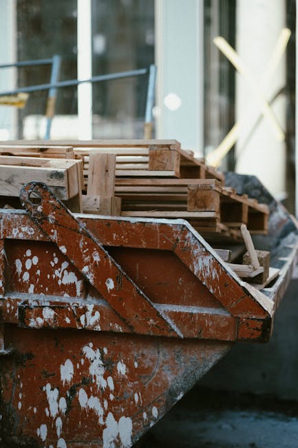 A close-up view of an orange-painted construction skip filled with wooden pallets, situated outdoors near a modern glass building. The skip's surface shows signs of wear and patchy white paint splatters, with the pallets stacked neatly inside, varying in size and construction, some with rough, unfinished edges and others with smoother surfaces. The pallets are made of light-colored, untreated timber with visible grain and texture, and are positioned horizontally and slightly leaning against each other. The background features blurred elements of the urban environment, including reflective glass windows and metallic railing supports, indicating a contemporary setting. The scene is illuminated by natural daylight, emphasizing the textures of the weathered skip and the raw timber of the pallets, creating a neutral, professional appearance aligned with on-site waste management operations handled by companies like Rubbish Removal Kennington, supporting alternative waste handling methods outside of traditional municipal refuse collection.