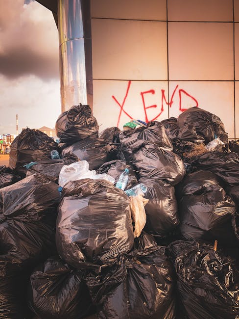 A large pile of black plastic rubbish bags, some torn open revealing plastic bottles, paper, and miscellaneous waste inside. The bags are stacked on the ground next to a modern building with a reflective metal column on the left and a tiled wall in the background. Red graffiti spelling 'XEND' is spray-painted on the wall behind the rubbish. The environment appears to be an urban setting, possibly on a pavement or waste collection area, with cloudy sky visible in the top left corner. The scene illustrates an instance of unmanaged waste accumulation, which is relevant to private rubbish removal services like those offered by Rubbish Removal Kennington, highlighting the need for professional, independent waste disposal solutions for such cluttered and unsanitary conditions.