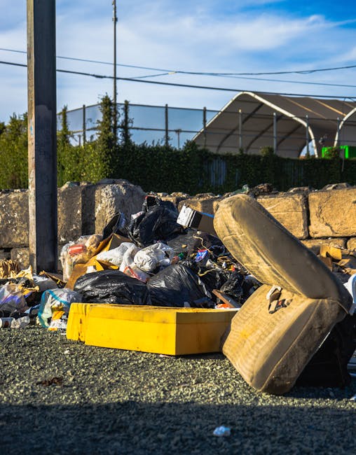 Three large black plastic rubbish bags filled with waste are positioned on the pavement near a black wooden fence, with a hedge of dark foliage behind them. The bags appear to be tightly packed and are leaning slightly, indicating they are ready for collection or disposal. The environment suggests an urban or residential street setting, with the waste bags placed close to the curb, possibly awaiting private rubbish removal services offered by companies such as Rubbish Removal Kennington. The scene is captured in natural lighting, with shadows cast by nearby objects, emphasizing the texture of the plastic bags, which have visible creases and some printed material on their surfaces. The overall composition highlights a typical waste management scenario involving on-site clearance of household waste, aligning with private rubbish disposal or alternate waste handling methods, rather than council-led collection.